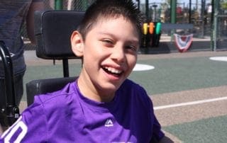 A young boy in a wheelchair smiling at a baseball field.